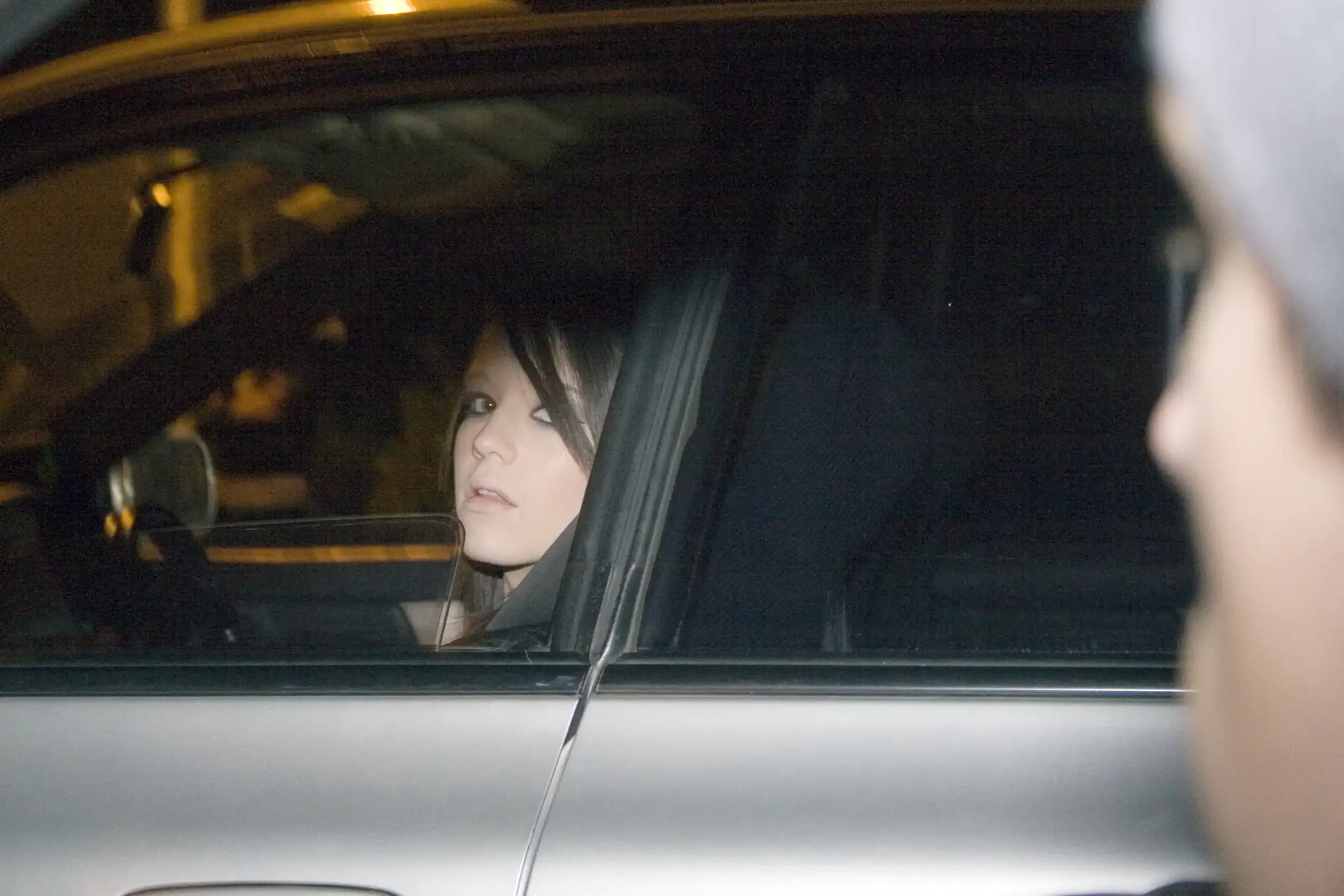 A young woman sits in the driver's seat of a parked car at night, watched through the window by a figure in the foreground.