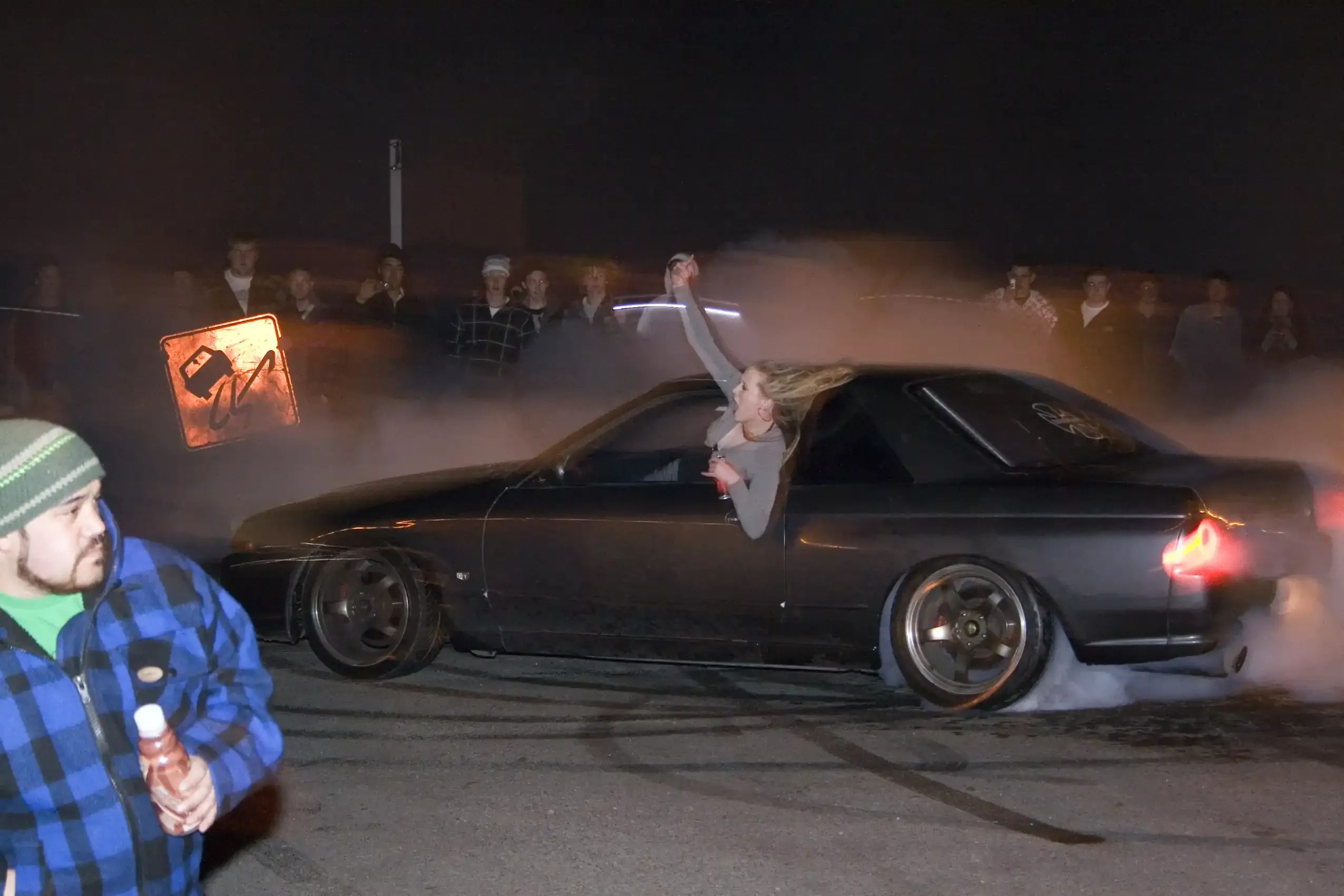 A car doing a smoky burnout at a night meet as a crowd watches.