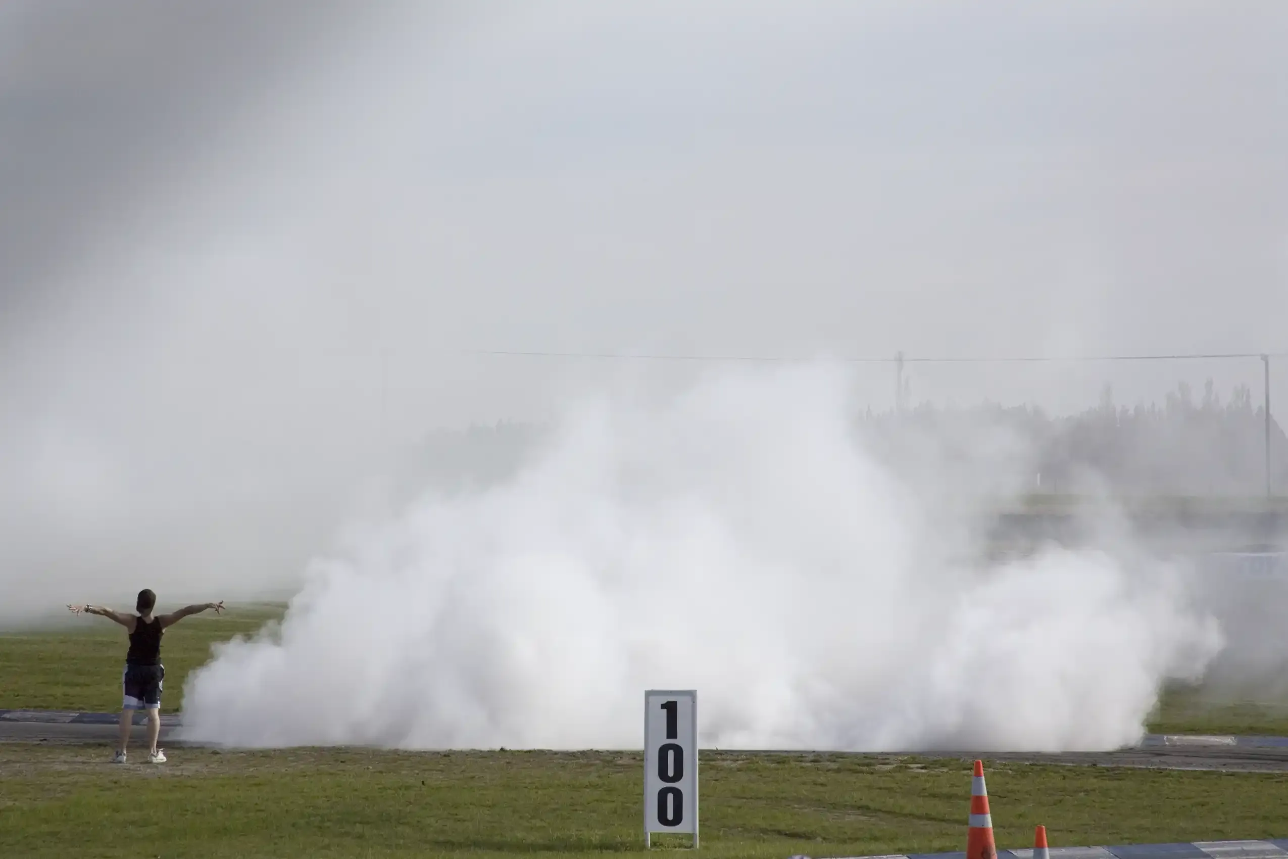 Daytime drag strip: a person stands arms-outstretched beside a '100' distance marker as a car vanishes into a vast cloud of tyre smoke.